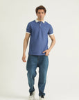 A man wearing a blue textured knitted cotton polo shirt with a collar, short sleeves, and a straight-cut hem with a small slit at each side. He is also wearing blue jeans and white sneakers.