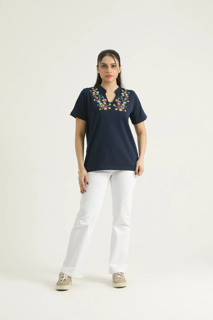 A woman standing against a white background wearing a navy blue knit top with a floral print and a V-neckline, paired with white pants and beige shoes.