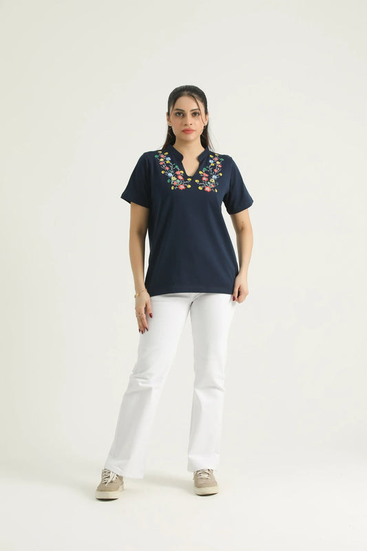 A woman standing against a white background wearing a navy blue knit top with a floral print and a V-neckline, paired with white pants and beige shoes.