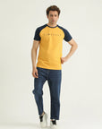 A man wearing a yellow and navy contrast raglan sleeve t-shirt with a round neckline and ribbed hem, paired with blue jeans and white sneakers.