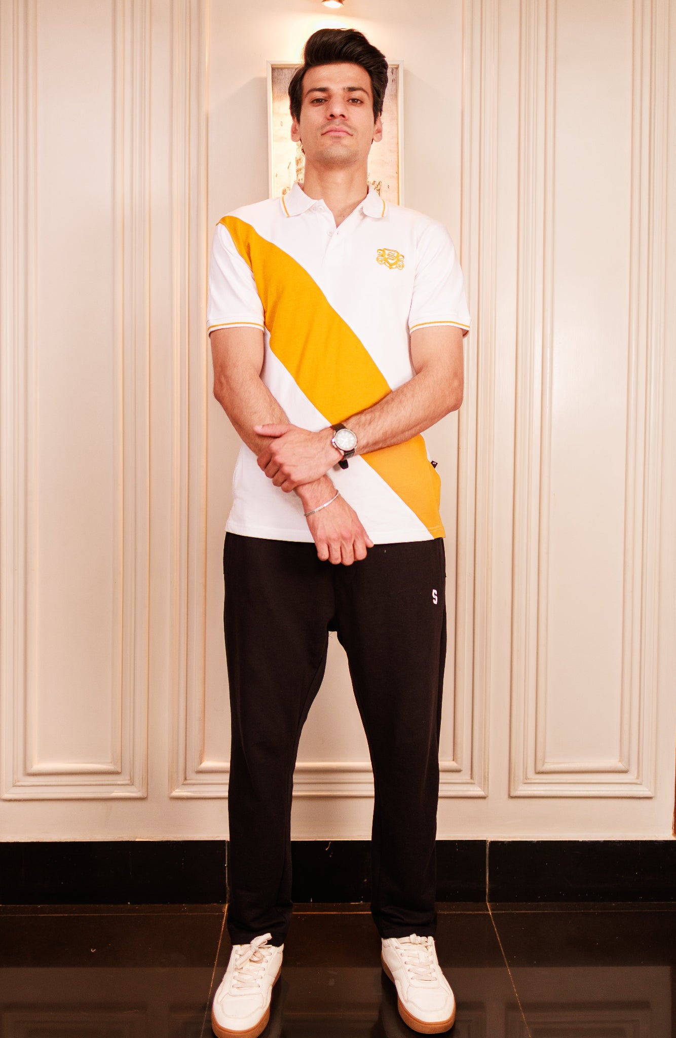 Man wearing a white polo shirt with a yellow diagonal stripe and black pants, standing against a light-colored paneled wall.