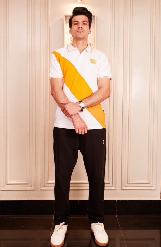 Man wearing a white polo shirt with a yellow diagonal stripe and black pants, standing against a light-colored paneled wall.