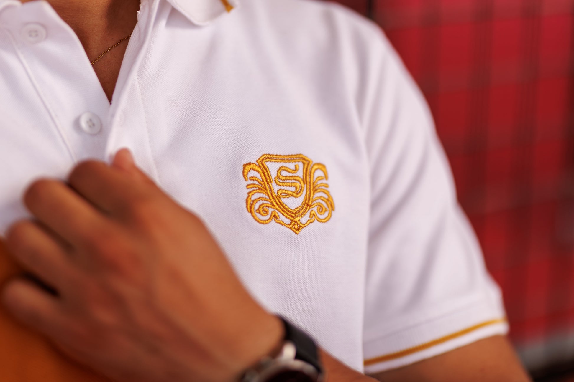 White polo shirt with gold emblem worn by a person, blurred red background