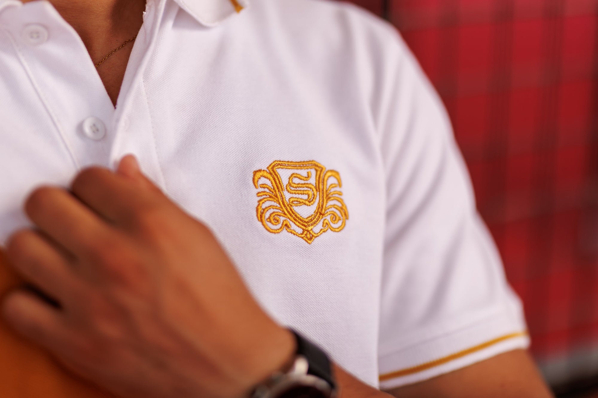 White polo shirt with gold emblem worn by a person, blurred red background