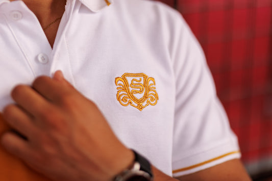 White polo shirt with gold emblem worn by a person, blurred red background