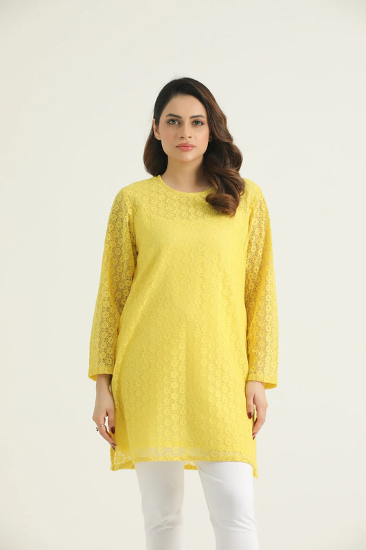 A woman wearing a bright marigold yellow mid-length top with long sleeves and floral lace details, paired with white pants.