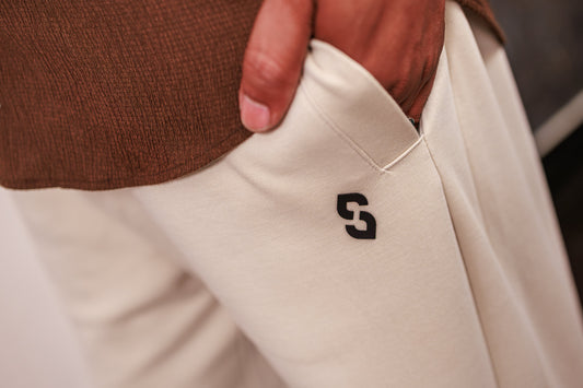 Close-up of beige pants with a logo on the pocket, worn by a person in a brown sweater.