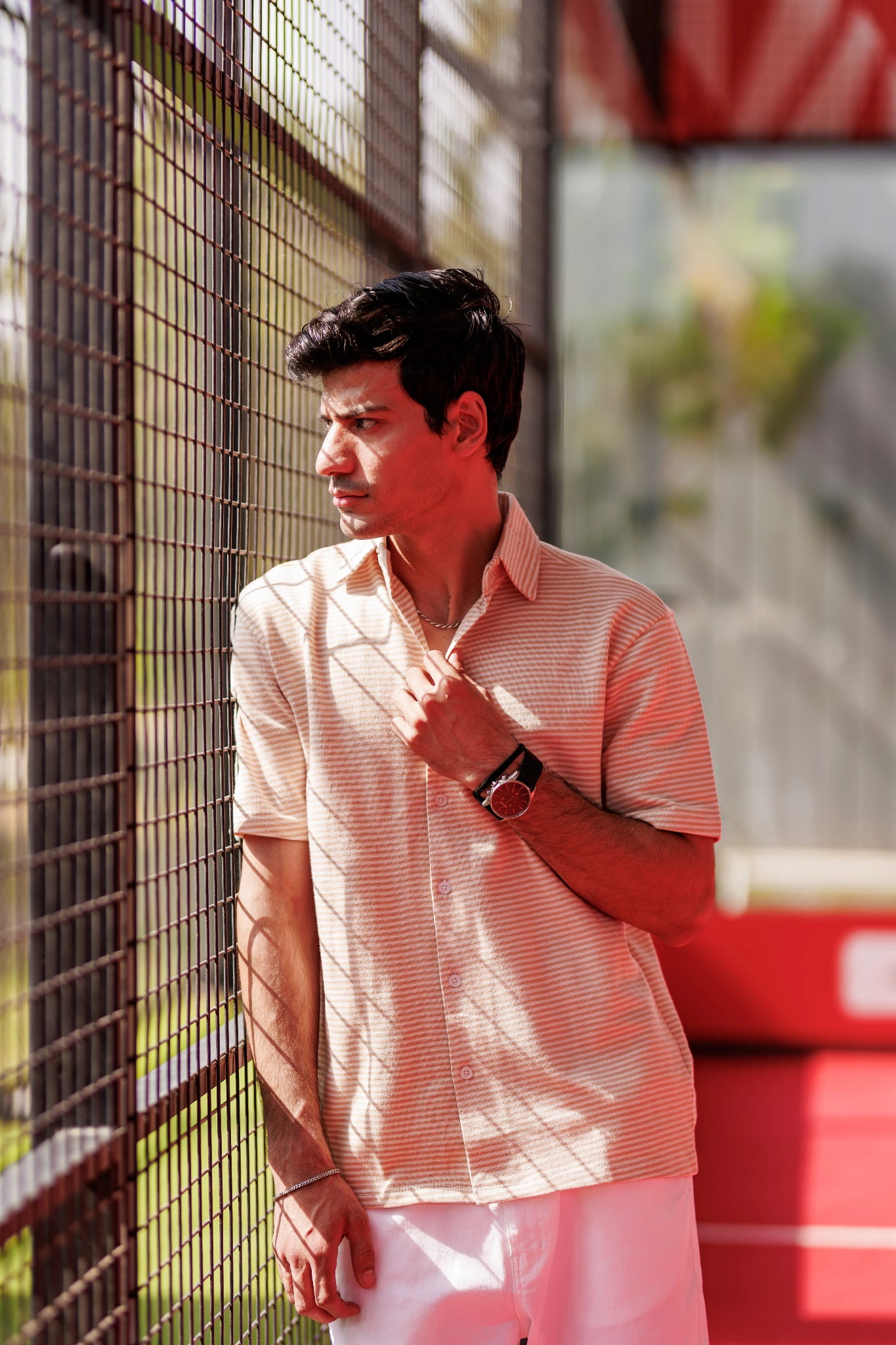 Man wearing a light pink shirt and white pants standing near a metal fence.
