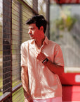 Man wearing a light pink shirt and white pants standing near a metal fence.