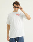 A man wearing a white loose fit printed t-shirt with red text on the front, paired with blue denim jeans.