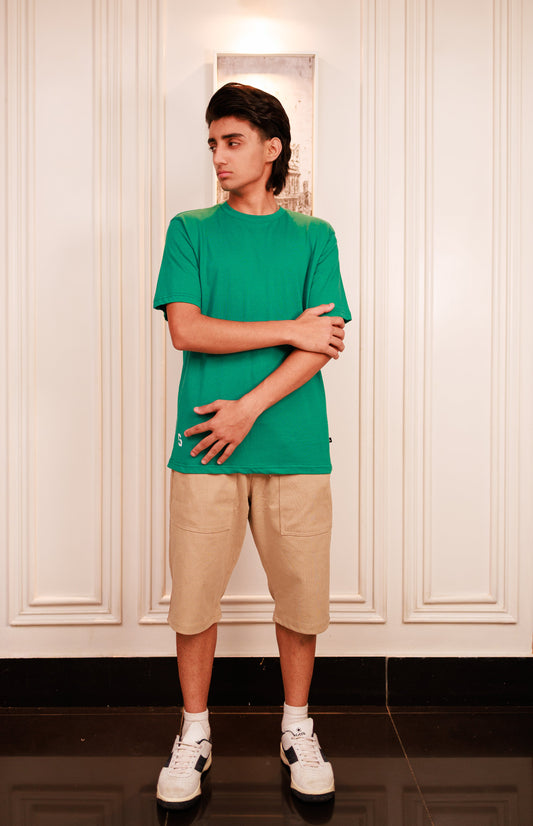 Person wearing a green t-shirt and beige shorts standing against a white paneled wall.