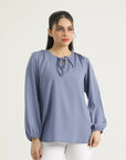 A woman modeling a long-sleeved, gathered-neck, dusty periwinkle blue blouse with a straight-cut silhouette.
